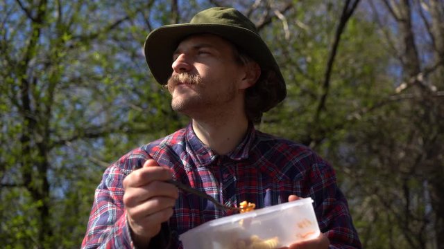 young handsome hipster millennial traveler hiker in checkered shirt and hat eats fast made pasta salad lunch in camping, person enjoys nature outside freedom holidays in forest