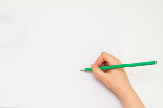 Top view of hand of kid ready to draw with green color pencil on white paper background. Copy space for text. Mockup.