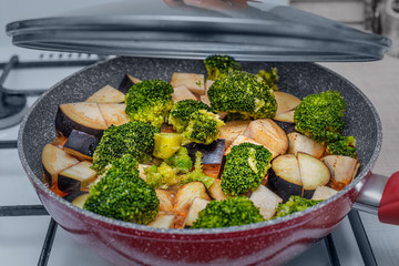 Cooking delicious and healthy vegetable vegetarian stew in a frying pan on a gas stove. The recipe includes eggplant, broccoli and other ingredients
