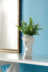 Subject shot of a white ceramic flower pot with a bouquet of artificial flowers. The pot made as an antique head is located on the white table against the blue wall with a mirror in a golden frame. 