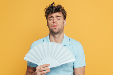 Man with closed eyes and hand fan suffering from heat isolated on yellow