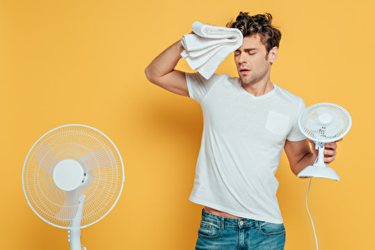 Man with towel, electric and desk fans suffering from heat on yellow