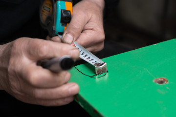 Fototapeta premium An old man in his workshop is making measurements with measuring tape and making marks with black pen on a peace of green woden plate