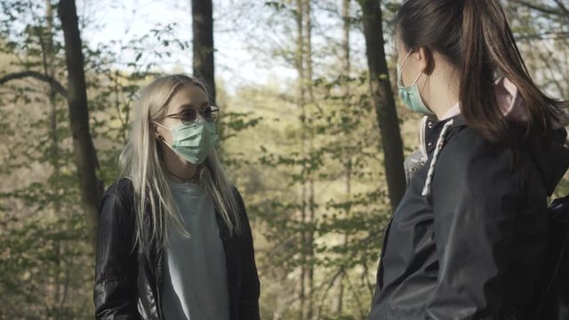 Portrait of blond young woman in face mask talking with friend in sunny park. Two millennial girls chatting outdoors on coronavirus lockdown. Covid-19 pandemic, quarantine lifestyle.