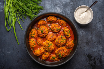 Homemade meatballs in tomato sauce in a saucepan against a dark background. Next to the saucer with sour cream and greens