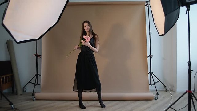 a tall girl with long hair moves on a beige background, is in a photo studio, softboxes stand nearby, the girl holds a flower in her hands, swirls, jumps and smiles
