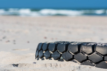 Big black rubber tire left on a sandy beach with blue sea and waves on background, environment pollution concept