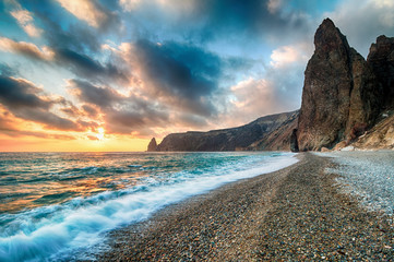 Bright sea sunset. The waves crash into the rock, lit by the warm sunset, sand and pebbles,...