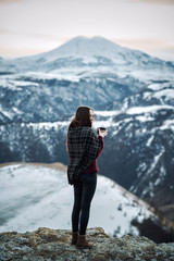 Fototapeta premium Young brunette girl drinks hot tea in the winter mountains of the Caucasus. The girl wrapped herself in a warm plaid and holds a mug of hot tea in the winter traveling in the mountains