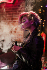 Fototapeta na wymiar a girl with bright colored purple pink hair in a leather jacket black t shirt jeans in a dark dark bar against the background of neon signs poses smoking vape