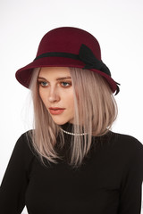 A blonde European lady with blunt bob haircut is wearing a burgundy cloche hat, a black polo-neck and a golden necklace with crystals. The hat is decorated with a wide black band and a bow. 