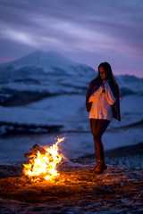 A young girl stands at sunset by the fire and looks at the flame. A woman wrapped in a warm plaid stands on a cliff and admires the flames of the fire. In the background snowy mountains and Elbrus