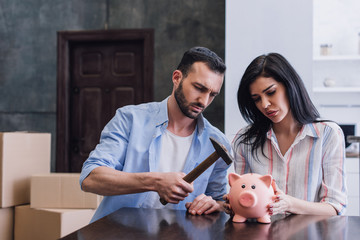 Woman with upset man holding hammer above piggy bank at table in room