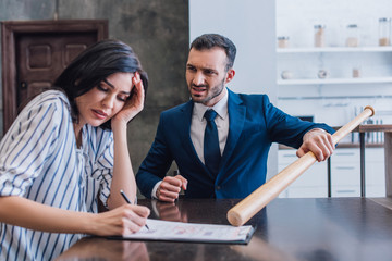 Scared woman writing in documents near angry collector with baseball bat at table in room
