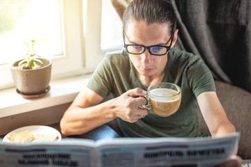 Fototapeta na wymiar Young handsome man reading interesting hot news articles in a newspaper and drinking Cup of cappuccino coffee