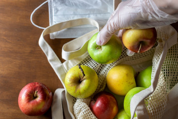 Hand in glove holds apple. Eco mesh bag with food supplies on a wooden background. Food delivery in eco-friendly packaging, zero waste.