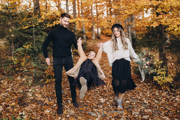 Stylish family in the autumn forest. A young guy and a girl are walking along a forest road among yellow leaves with their daughter.