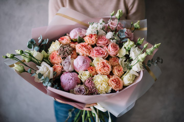 Very nice young woman holding big beautiful blossoming bouquet of fresh peony, roses, eustoma, carnations, eucalyptus, flowers in pink, yellow and white colors on the grey wall background