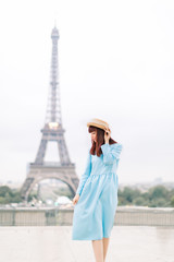 Fototapeta na wymiar Romantic beautiful girl portrait with tour eiffel on the background. Pretty tender young lady in blue dress and hat touching her red hair and walking in Paris