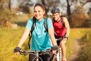 Obraz premium Beautiful happy young Caucasian woman riding mountain bike over a meadow in the early morning and her boyfriend is following. Healthy and active sporty lifestyle