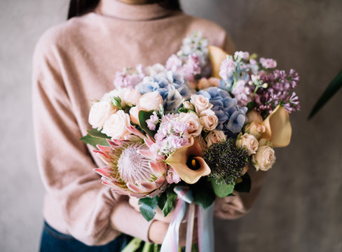 Very nice young woman holding big beautiful blossoming bouquet of fresh hydrangea, nutan protea, lilac, matthiola, roses, calla lilies flowers in blue, pink, cream and lilac colors 