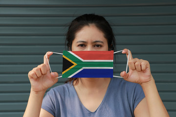 A woman with South Africa flag on hygienic mask in her hand and lifted up the front face on dark green background. Tiny Particle or virus corona or Covid 19 protection.