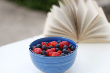 Bowl of strawberries and blueberries and open book in a garden. Selective focus.