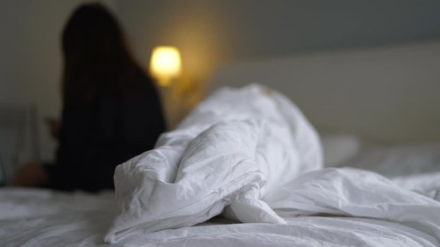 Unmade bed with crumpled bed sheet, blanket and pillows after sleep waking up in the morning, woman on bed using mobile phone, shallow depth of field.