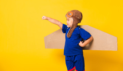 Happy Asian handsome funny child or kid little boy smile wear pilot hat play and goggles raise hand up with toy cardboard airplane wings flying, studio shot isolated yellow background, Startup freedom