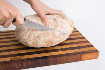 Hands of person cutting round loaf of freshly baked crusty bread on chopping board. Isolated object on white background. Healthy food for breakfast concept