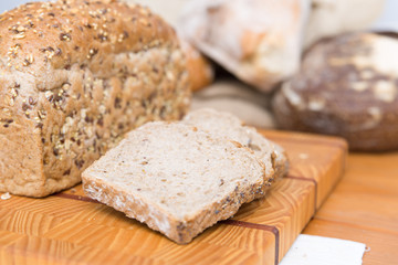 Freshly baked wholegrain bread slices for breakfast on chopping board. Closeup shot. Heap of loafs and buns in background. Bakery or healthy eating concept