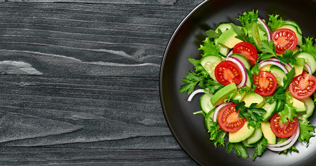 Fresh salad with cherry tomatoes, avocado, cucumeber on dish. Healthy vegetarian meal concept. Summer mixed leaves, tomatoes and vegetable salad on black wood, top view.