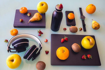 Table with different black objects, laptop, watch, bottle, phone, headphones and a book, organized. And different fruits such as oranges, pears, apples etc decorating it.