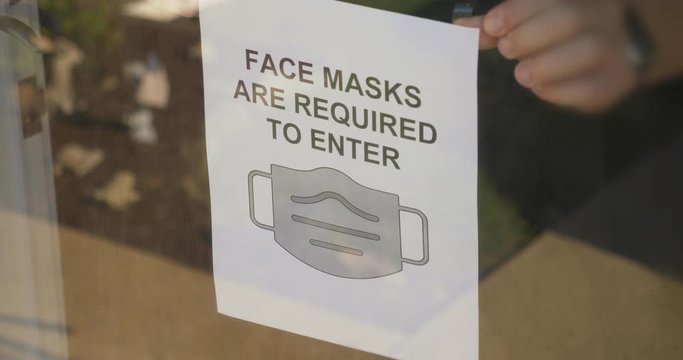 A store owner puts a COVID-19 face mask warning sign on the front door. Wearing face masks was often required when entering a business during the coronavirus pandemic of 2020.  	