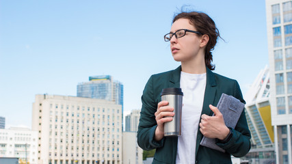 Serious millennial female office worker standing on city street holding tablet computer and coffee....