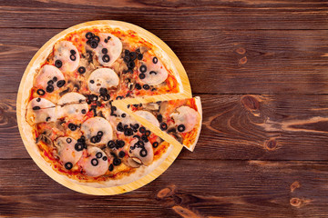 Pizza with ham, mozzarella, mushrooms and olives, with a slice slightly removed on a wood platter which is on wooden rustic table, top view and copy space
