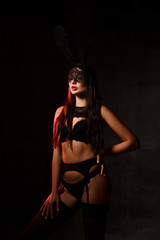 Brunette woman in black lace sexy lingerie, garter belt, stockings and mask stands in twilight against black background