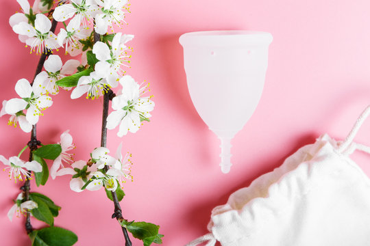 Eco white menstrual cup and cotton bag on a pink background, spring cherry tree branches with white flowers. Ecology and recycling concept, zero waste. Women's hygiene, menstruation, critical days.