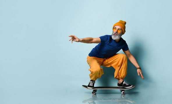 Bearded Elderly Man In T-shirt, Sunglasses, Orange Pants, Hat, Gumshoes. Riding Black Skateboard, Posing On Blue Background