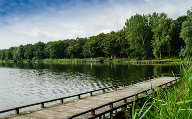 Fototapeta na wymiar Netherlands, Zeeland region. August 2019. The beautiful pond of a campsite: the wooden pier goes into the water, in the background the forest runs alongside the shore.