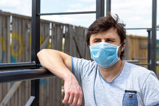 Man in protective blue medical mask standing on the outdoor sports ground, resting his hand on the bars. Male in a respirator to protect against infection with virus during training in quarantine