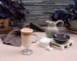 A popular Korean drink during self-isolation is Dalgona coffee. A delicious modern cold beverage with chocolate. Creamer, napkin is on the table. On a background is potted plants. Vintage still life.