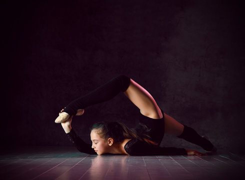 Young girl gymnast in black sport body and uppers lying on floor on neck with streched back and body and making gimnastic pose