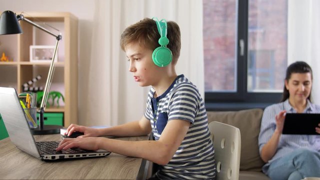 children, education and distant learning concept - student boy in headphones with laptop computer and mother with tablet pc at home