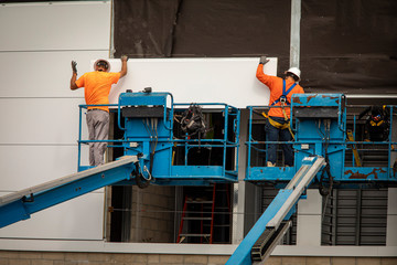 Two construction workers wearing hardhats on a blue manlift hanging section of drywall paneling on prefab building