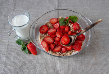 Muesli with yogurt and strawberries in a glass bowl, next to a glass of milk and a wooden spoon.