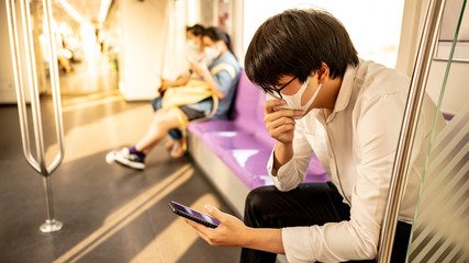 Fototapeta na wymiar Asian man wearing surgical face mask using smartphone on skytrain or urban train. Coronavirus (COVID-19) outbreak prevention in public transportation. Health awareness for pandemic protection
