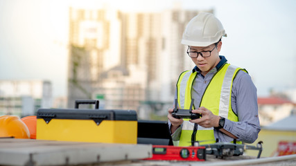 Asian engineer man working with drone, laptop and working tools at construction site. Male worker using unmanned aerial vehicle (UAV) for land and building site survey in civil engineering project.