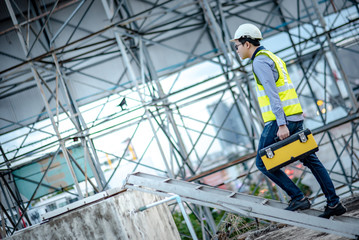 Asian maintenance worker man with safety helmet and green vest carrying tool box climbing on aluminium ladder at construction site. Civil engineering, Architecture builder and building service concept