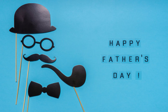 Various black photo booth props: cylinder hat, glasses, moustache, smoking pipe, bow tie on blue background. Greeting card, text HAPPY FATHER'S DAY. Creative composition in minimal style
